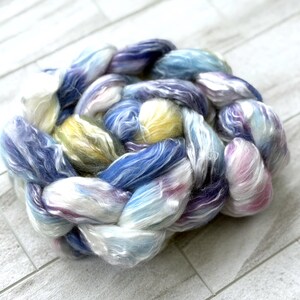 Op de afbeelding: Een close-up van een gevlochten streng handgeverfd garen. Het garen is een mix van blauw, paars, geel en wit. De vezels zijn zacht en pluizig, perfect voor brei- of haakprojecten.