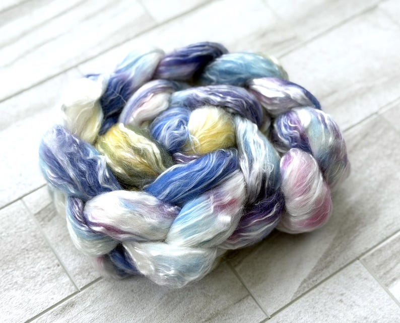 Op de afbeelding: Een close-up van een gevlochten streng handgeverfd garen. Het garen is een mix van blauw, paars, geel en wit. De vezels zijn zacht en pluizig, perfect voor brei- of haakprojecten.