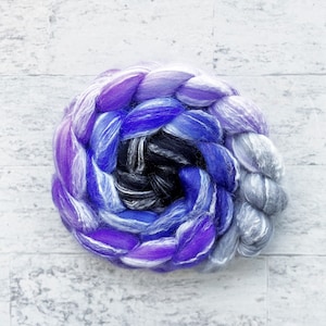 May include: A close-up of a hand-spun yarn ball in shades of purple, blue, and gray. The yarn is arranged in a spiral pattern, creating a visually appealing and textured effect.