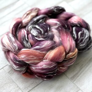 May include: A close-up of a skein of hand-dyed fiber in shades of pink, purple, and white. The fiber is arranged in a braided pattern, with the colors blending together. The fiber is soft and fluffy, perfect for spinning or felting.
