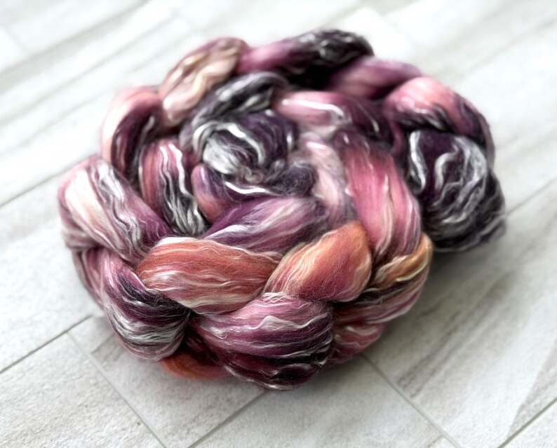 May include: A close-up of a skein of hand-dyed fiber in shades of pink, purple, and white. The fiber is arranged in a braided pattern, with the colors blending together. The fiber is soft and fluffy, perfect for spinning or felting.