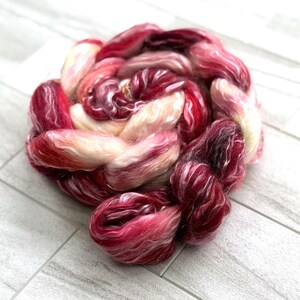 May include: A close-up of a skein of hand-dyed fiber in shades of red, pink, and cream. The fiber is arranged in a circular shape, with the colors blending together. The texture of the fiber is soft and fluffy.