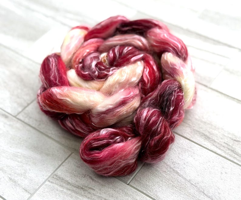 May include: A close-up of a skein of hand-dyed fiber in shades of red, pink, and cream. The fiber is arranged in a circular shape, with the colors blending together. The texture of the fiber is soft and fluffy.