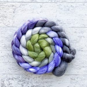 May include: A hand-spun yarn braid in shades of purple, green, gray, and white. The yarn is arranged in a spiral pattern.