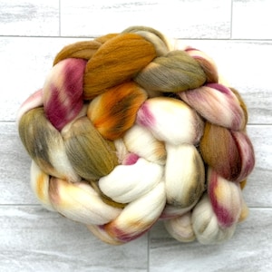 May include: A braided skein of colorful wool roving. The roving features a blend of colors, including white, yellow, orange, green, and shades of pink and brown. The wool is arranged in a thick, textured braid, ideal for fiber art projects.
