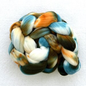 Könnte beinhalten: Ein dickes, geflochtenes Bündel aus Kammzugwolle mit einer bunten Farbpalette aus Weiß, Himmelblau, tiefem Waldgrün und gebranntem Orange. Die weiche, flauschige Textur ist in den ineinander verschlungenen Fasersträngen deutlich erkennbar.