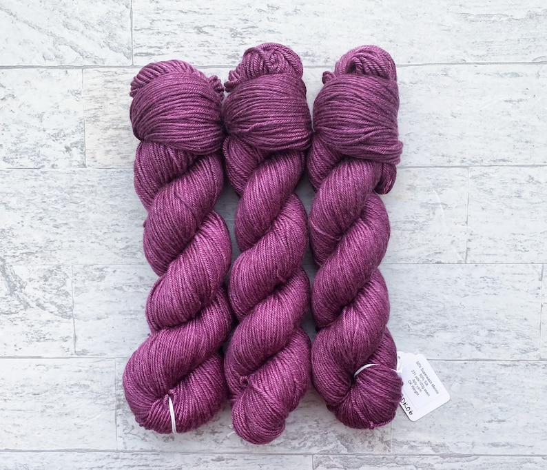 May include: Three skeins of deep purple yarn, twisted together. The yarn is soft and has a subtle sheen. The skeins are labeled with the text "100% Superwash Merino Wool, 117 yards, 100g, 4 ply, DK"