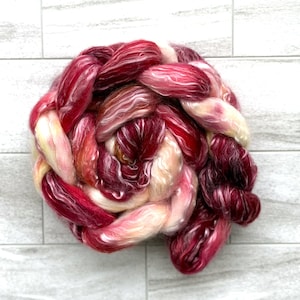May include: A skein of hand-dyed yarn in shades of red, burgundy, and cream. The yarn is arranged in a braided or twisted form, showcasing the color variations. The fiber appears soft and luxurious, suitable for knitting or crochet projects.