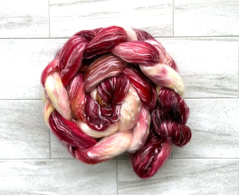 May include: A skein of hand-dyed yarn in shades of red, burgundy, and cream. The yarn is arranged in a braided or twisted form, showcasing the color variations. The fiber appears soft and luxurious, suitable for knitting or crochet projects.
