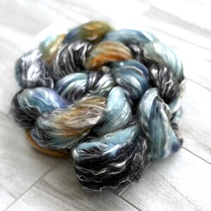 May include: A ball of hand-dyed yarn in shades of blue, grey, and gold. The yarn has a soft, textured appearance, with colours blended to create a marbled effect. Suitable for knitting, crochet, or other fibre arts projects. The yarn is a blend of various fibres.