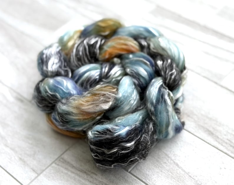 May include: A ball of hand-dyed yarn in shades of blue, grey, and gold. The yarn has a soft, textured appearance, with colours blended to create a marbled effect. Suitable for knitting, crochet, or other fibre arts projects. The yarn is a blend of various fibres.