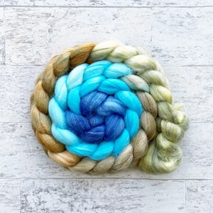 May include: A braided bundle of yarn in shades of green, blue, and brown. The yarn is arranged in a spiral pattern, with the green yarn on the outside, the blue yarn in the middle, and the brown yarn on the inside.