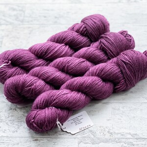 May include: Three skeins of deep purple yarn, each skein is wound into a ball shape. The yarn is labeled "50% Superwash Merino 25% Silk 25% Nylon 437 yards / 400 meters DK weight"