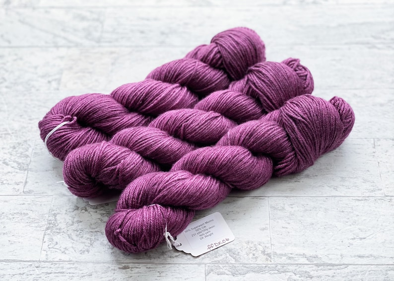May include: Three skeins of deep purple yarn, each skein is wound into a ball shape. The yarn is labeled "50% Superwash Merino 25% Silk 25% Nylon 437 yards / 400 meters DK weight"