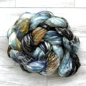 May include: A braided skein of yarn in shades of blue, green, brown, and black. The yarn has a soft, textured appearance, suitable for knitting or other fibre art projects. The skein is displayed against a white background.