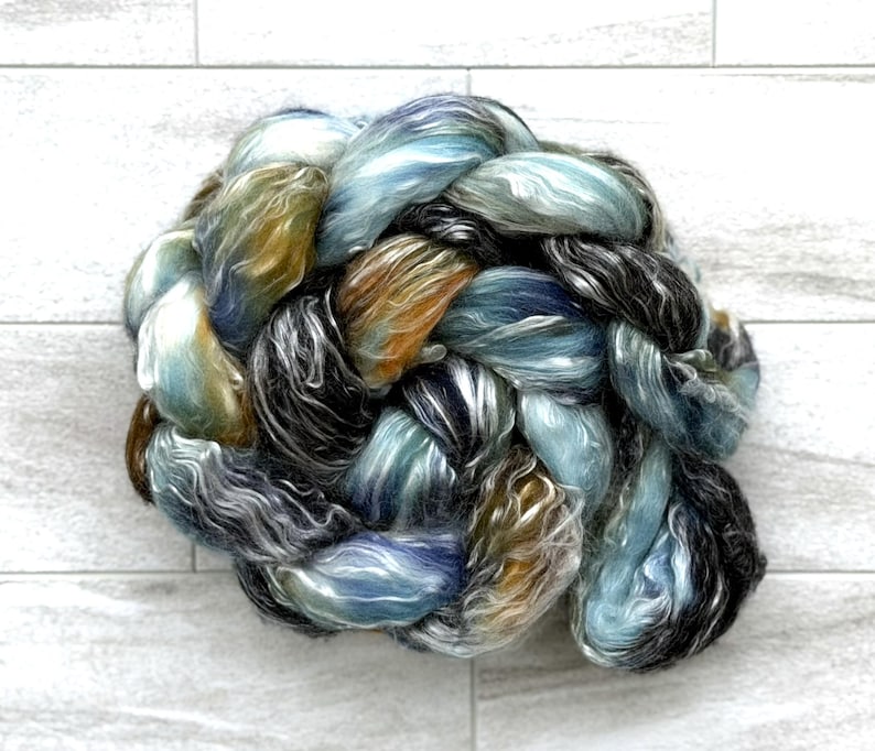 May include: A braided skein of yarn in shades of blue, green, brown, and black. The yarn has a soft, textured appearance, suitable for knitting or other fibre art projects. The skein is displayed against a white background.