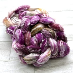 Puede incluir: Un ovillo trenzado de hilo te&ntilde;ido a mano en tonos morado, blanco y marr&oacute;n. El hilo tiene una textura suave y esponjosa y est&aacute; dispuesto en forma circular. Los colores se mezclan, creando un producto de fibra visualmente atractivo y texturizado.