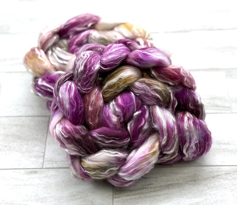 Puede incluir: Un ovillo trenzado de hilo te&ntilde;ido a mano en tonos morado, blanco y marr&oacute;n. El hilo tiene una textura suave y esponjosa y est&aacute; dispuesto en forma circular. Los colores se mezclan, creando un producto de fibra visualmente atractivo y texturizado.