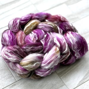 Puede incluir: Un ovillo trenzado de fibra te&ntilde;ida a mano en tonos morados, blancos y marrones. La fibra est&aacute; dispuesta en forma circular, con los colores mezcl&aacute;ndose para crear una textura visualmente atractiva. Ideal para tejer, tejer o otras artes de la fibra.