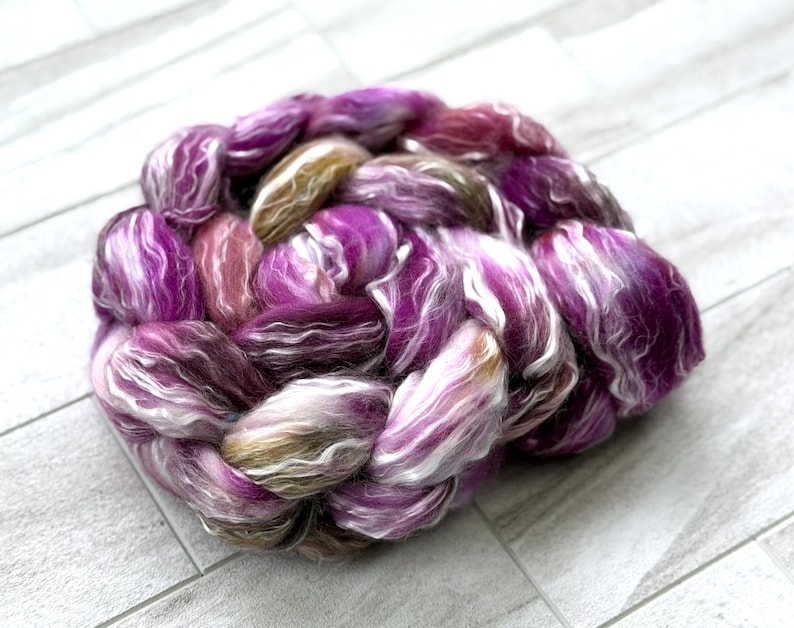 Puede incluir: Un ovillo trenzado de fibra te&ntilde;ida a mano en tonos morados, blancos y marrones. La fibra est&aacute; dispuesta en forma circular, con los colores mezcl&aacute;ndose para crear una textura visualmente atractiva. Ideal para tejer, tejer o otras artes de la fibra.