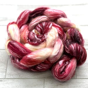 May include: A tightly wound braid of hand-dyed fiber in shades of red, pink, and cream. The fiber appears soft and fluffy, with a subtle sheen. Ideal for spinning, weaving, or other fiber arts projects.