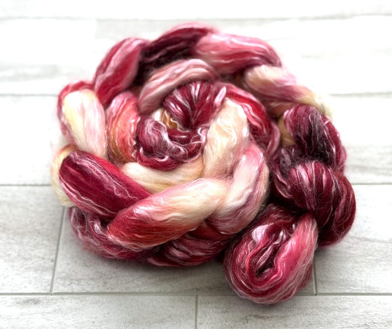 May include: A tightly wound braid of hand-dyed fiber in shades of red, pink, and cream. The fiber appears soft and fluffy, with a subtle sheen. Ideal for spinning, weaving, or other fiber arts projects.