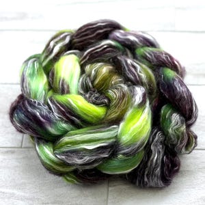 May include: A braided bundle of colorful fiber for crafting. The fiber is a mix of green, purple, white, and brown. The colors are blended together to create a unique and textured look. This fiber is ideal for spinning, weaving, or other fiber art projects.