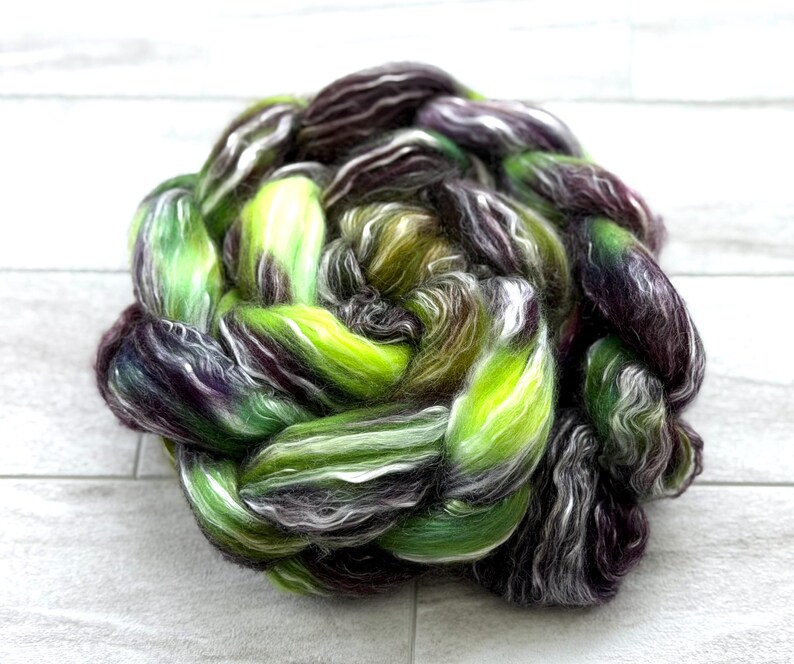 May include: A braided bundle of colorful fiber for crafting. The fiber is a mix of green, purple, white, and brown. The colors are blended together to create a unique and textured look. This fiber is ideal for spinning, weaving, or other fiber art projects.