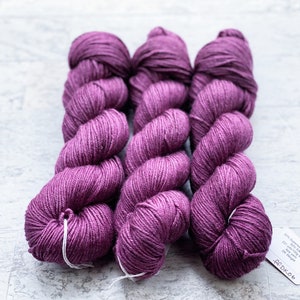May include: Three skeins of purple yarn, each wound into a ball shape. The yarn is a medium weight and has a subtle sheen. The label on the bottom skein reads "60% Superwash Merino 100g 325 yds 4 ply DK Weight".