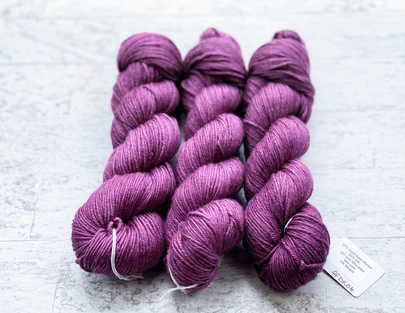 May include: Three skeins of purple yarn, each wound into a ball shape. The yarn is a medium weight and has a subtle sheen. The label on the bottom skein reads "60% Superwash Merino 100g 325 yds 4 ply DK Weight".