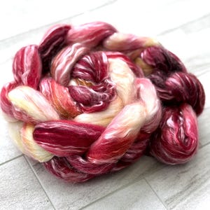 May include: A close-up of a braided bundle of colorful fiber. The fiber is a mix of red, pink, and cream colors. The fiber is soft and fluffy, and it appears to be hand-dyed. The bundle is sitting on a white wooden surface.