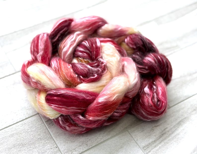 May include: A close-up of a braided bundle of colorful fiber. The fiber is a mix of red, pink, and cream colors. The fiber is soft and fluffy, and it appears to be hand-dyed. The bundle is sitting on a white wooden surface.