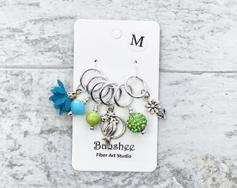 Puede incluir: Conjunto de cinco marcadores de punto de plata con abalorios verdes y azules. Los abalorios incluyen un pájaro, una flor, una cuenta y una bola verde brillante. La tarjeta dice "M" y "Balshee Fiber Art Studio".