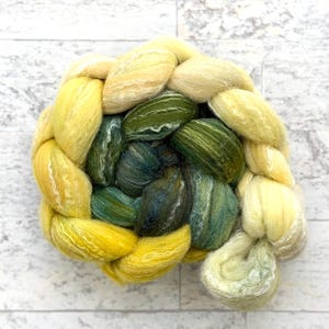 May include: A braided skein of hand-dyed yarn in shades of yellow, green, and teal. The yarn is arranged in a circular pattern, showcasing the color variations and texture. Ideal for knitting, crochet, or other fiber arts projects.