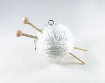 Puede incluir: Una bola de hilo blanco con dos agujas de tejer de madera y un porta anillos de plata.