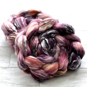 May include: A large, braided skein of hand-dyed yarn in shades of pink, purple, and peach. The yarn is thick and textured, with a mix of solid and variegated colors. The skein is sitting on a white, wooden surface.