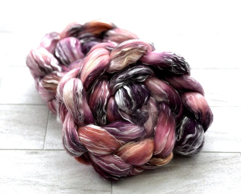 May include: A large, braided skein of hand-dyed yarn in shades of pink, purple, and peach. The yarn is thick and textured, with a mix of solid and variegated colors. The skein is sitting on a white, wooden surface.