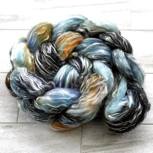 May include: A close-up of a braided skein of hand-dyed yarn. The yarn features a blend of colours, including blues, greens, oranges, and black, creating a textured and artistic appearance. The yarn is likely used for knitting or other fibre crafts.