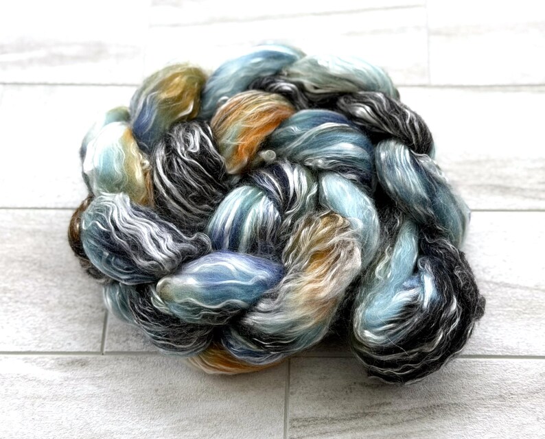 May include: A close-up of a braided skein of hand-dyed yarn. The yarn features a blend of colours, including blues, greens, oranges, and black, creating a textured and artistic appearance. The yarn is likely used for knitting or other fibre crafts.