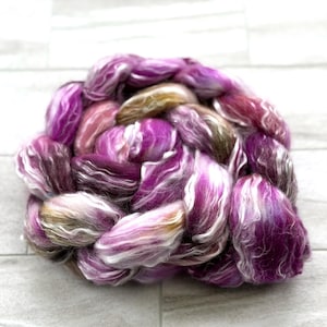 Puede incluir: Primer plano de una madeja de hilo te&ntilde;ido a mano en tonos morado, rosa, blanco y dorado. El hilo est&aacute; bien enrollado, mostrando las variaciones de textura y color. El hilo est&aacute; sobre una superficie gris claro.