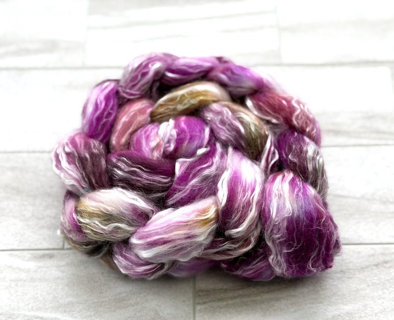 Puede incluir: Primer plano de una madeja de hilo te&ntilde;ido a mano en tonos morado, rosa, blanco y dorado. El hilo est&aacute; bien enrollado, mostrando las variaciones de textura y color. El hilo est&aacute; sobre una superficie gris claro.