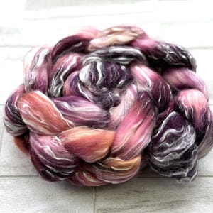 May include: A close-up of a braid of colorful fiber in shades of purple, pink, orange, and white. The fiber is soft and fluffy, with a slight sheen. The braid is tightly woven and appears to be ready for spinning or other fiber arts projects.