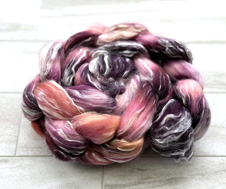 May include: A close-up of a braid of colorful fiber in shades of purple, pink, orange, and white. The fiber is soft and fluffy, with a slight sheen. The braid is tightly woven and appears to be ready for spinning or other fiber arts projects.