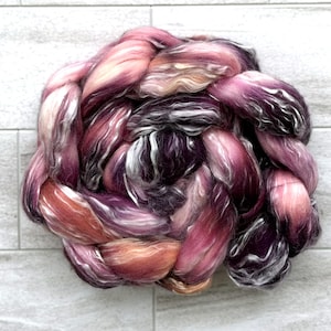 May include: A braided bundle of colorful fiber in shades of pink, purple, and orange. The fiber has a soft, textured appearance, with strands of white and dark purple woven throughout. The bundle is arranged in a circular shape, with the strands overlapping.