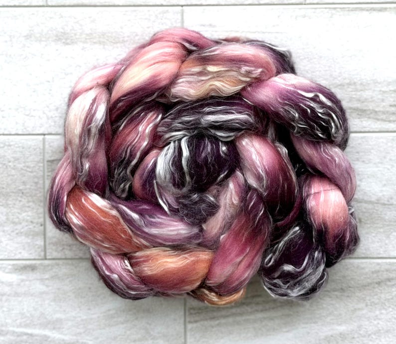 May include: A braided bundle of colorful fiber in shades of pink, purple, and orange. The fiber has a soft, textured appearance, with strands of white and dark purple woven throughout. The bundle is arranged in a circular shape, with the strands overlapping.