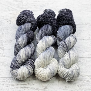 Puede incluir: Tres ovillos de hilo en un degradado de color negro y blanco. El hilo es de peso fino y tiene un brillo sutil.