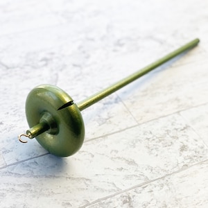 May include: A green wooden spinning wheel with a round, flat base and a long, thin spindle. The wheel is made of wood and has a smooth, polished finish.