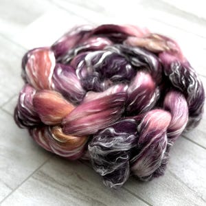 May include: A braided bundle of fiber in shades of pink, purple, and peach. The fiber has a soft, textured appearance, with strands of white and silver. The bundle is arranged in a circular shape, suitable for spinning or other fiber arts.