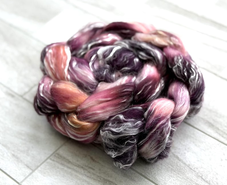May include: A braided bundle of fiber in shades of pink, purple, and peach. The fiber has a soft, textured appearance, with strands of white and silver. The bundle is arranged in a circular shape, suitable for spinning or other fiber arts.