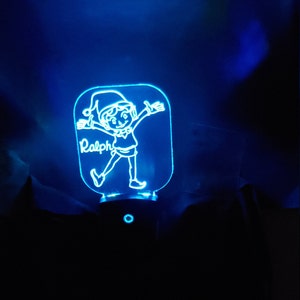 May include: A blue LED night light featuring a cartoon elf with outstretched arms. The name "Ralph" is inscribed on the light. The night light has a rectangular shape with rounded corners and a small base.